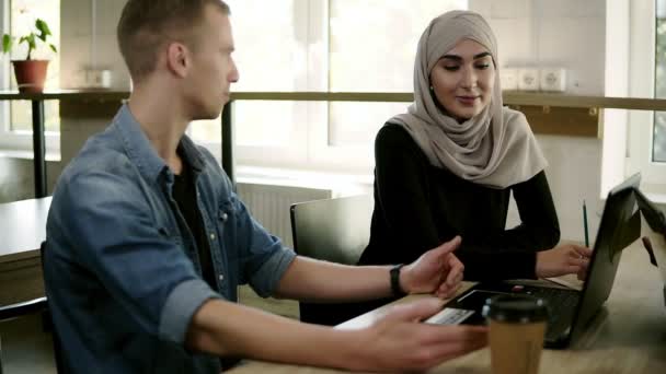 Запись многорасовых деловых встреч в современном светлом офисе за деревянным столом. Молодой человек в джинсовой рубашке объясняет проекты или состояние улыбающейся мусульманке в бежевом. — стоковое видео