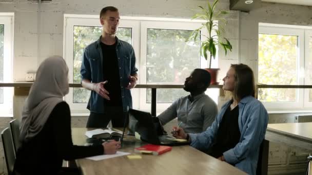 O jovem caucasiano no casual dá a explicação do projeto aos seus colegas ou parceiros. Equipe multirracial diversa sentada no espaço de trabalho moderno ouvindo palestrante apresentando novo projeto no escritório — Vídeo de Stock