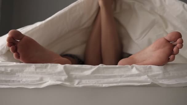 Close up of young beautiful and loving couple lying on the bed. Their feet under blanket while wake up in bed in morning. Woman is lying on man, her feet between his — Stock Video