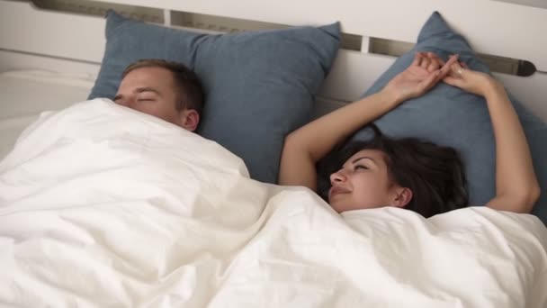 Joven mujer morena sonriente despertando feliz después de un sueño saludable estirándose en la cama cómoda con el hombre dormido cerca, chica refrescada disfruta de un agradable despertar, buenos días en el acogedor dormitorio loft y — Vídeos de Stock