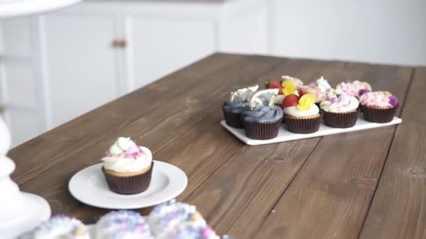 Footage of decorated pastry on the wooden table in studio kitchen. Cupcake with different colors mousse cream pink, blue, yellow with fruits and decoration in top. Tasty colorful cake on the stand — Stock Video