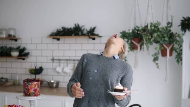 Neşeli mutlu kadın mutfağa üzerinde duran küçük lokum atmak ve ağzını ile yakalamak. Kısa gri saçlı kadın onun elinde geç üzerinde cupcake ile modern parlak mutfak ayakta. Sahip — Stok video