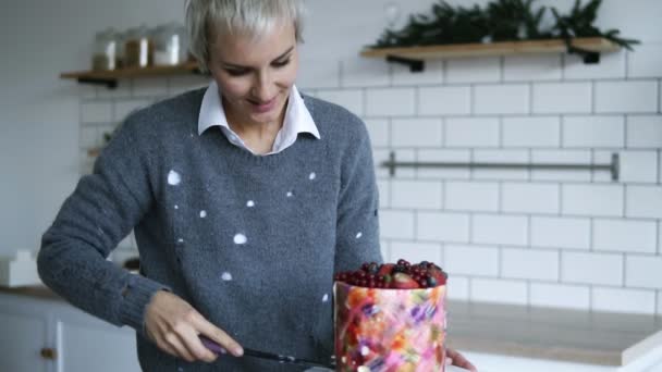Szürke hajú nő levágja egy késsel egy kis darab egy gyönyörű tortát, a desszert díszített bogyók a tetején, ő hozza, egy tányérra. Modern, fehér konyha — Stock videók