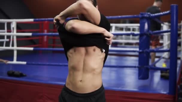 Tall handsome man undressing, taking off his black T shirt. Blue boxing ring at gym is in the background, revealing muscular naked torso in gym shot — Stock Video