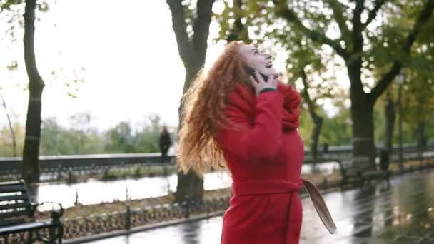 Jeune belle femme parlant sur son téléphone portable sur un fond. Fille excitée, heureuse aux cheveux bouclés rouges marchant dans le parc d'automne, tourbillonnant de plaisir. Femme en manteau rouge utilisant son smartphone. femme — Video