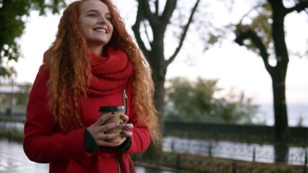 Šťastný, vzrušená žena v červeném kabátě chůzi s s sebou pití na ulici v deštivý podzim. Nádherná mladá dívka s dlouhými kudrnatými vlasy s úsměvem, se těší její čas venku — Stock video