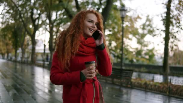 Молодая серьезная девушка разговаривает по мобильному телефону во время прогулки по осеннему парку. Кудрявая женщина в красном пальто пользуется смартфоном и держит бумажную чашку с кофе. Женщина ходит и разговаривает — стоковое видео