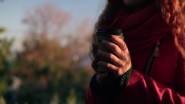 Wanita muda bergaya ceria bermantel merah syal berdiri di luar rumah sambil minum kopi di bawah sinar matahari di taman musim gugur. Gadis tersenyum menikmati hari-harinya. Uraikan cuplikan — Stok Video