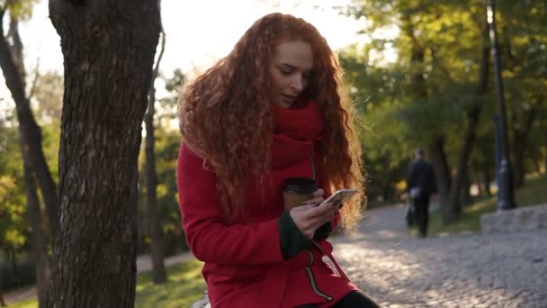 妇女卷曲的红色头发在红色外套和围巾站立在城市公园 使用手机和喝咖啡从科尔顿公园 — 图库视频影像