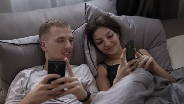 年轻的已婚夫妇躺在床上 互相分享手机上的搞笑照片 使用手机 快乐的生活方式 — 图库视频影像