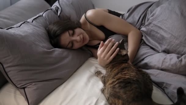 Mascotas, comodidad, descanso y concepto de la gente - mujer joven feliz con gato acostado en la cama en casa. Morena chica relajado acaricia su gato en la cama — Vídeo de stock