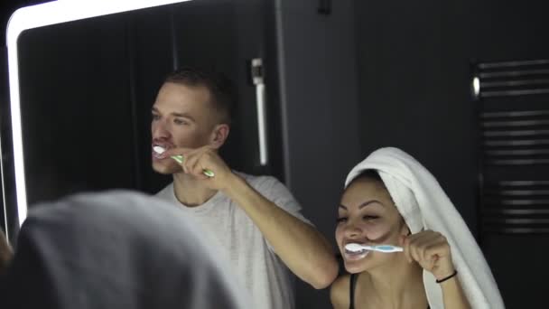 Joven pareja amorosa mirando en el espejo mientras se cepilla los dientes en el interior. Dama con toalla de baño en la cabeza — Vídeos de Stock
