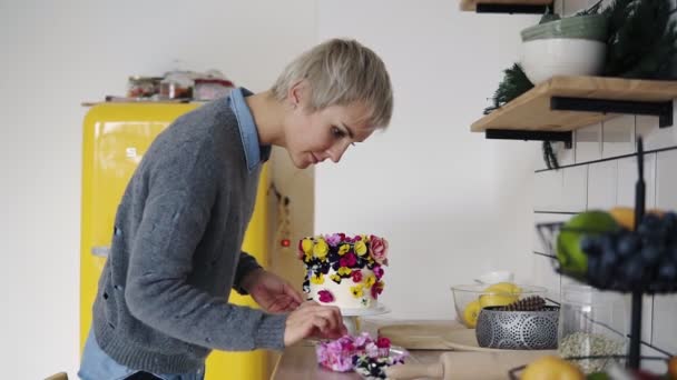 プロの菓子職人の女性は、白いモダンなキッチン スタジオの花とケーキを飾る。ショートヘアの女性シェフは、結婚式や誕生日のケーキ、食べられる新鮮な花になります。側からの映像 — ストック動画