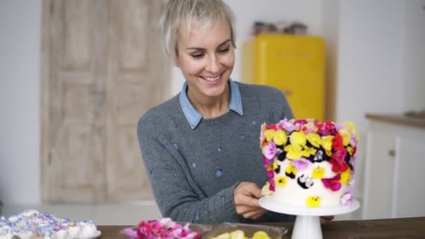 Χαμογελαστό γυναίκα σε γκρι πουλόβερ διακοσμεί τούρτα με λουλούδια στο λευκό σύγχρονη κουζίνα. Στενογραφία θηλυκό σεφ κάνει ένα γάμο ή γενέθλια τούρτα με λουλούδια φρέσκα, φαγώσιμο, η επιλογή των καλύτερων λουλουδιών για — Αρχείο Βίντεο