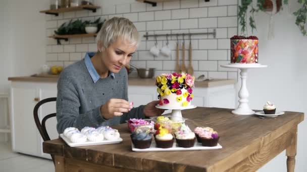 Konfektyr. Kocken dekorerar en order för en semester. Kvinna med grå färgat hår dekorerar tårtan med små blommor på beställning. Muffins på ett träbord på förgrunden — Stockvideo