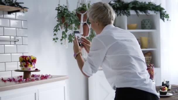 Blog de confitería. Mujer de camisa blanca fotografiando tarta decorada con flores. Pastelero decorado la ke pastel y luego toma fotos en el teléfono inteligente — Vídeos de Stock