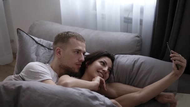 Утреннее селфи супружеской пары. Счастливая молодая пара лежит на кровати и делает автопортрет на смартфоне. Мужчина целует и обнимает жену, пока она снимает фильм. — стоковое видео