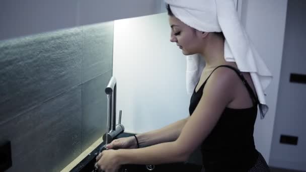 Mooie jonge lachende vrouw met witte badhanddoek op haar hoofd wassen een glas in de keuken. De routine van de ochtend na een douche. Zijaanzicht — Stockvideo