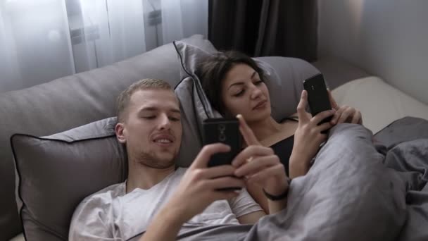 Молодая супружеская пара, лежа в постели и улыбаясь друг другу, пользуется мобильным телефоном. Счастливый образ жизни, пара, постель, хорошее настроение. Лежа на серых простынях, домашняя жизнь — стоковое видео