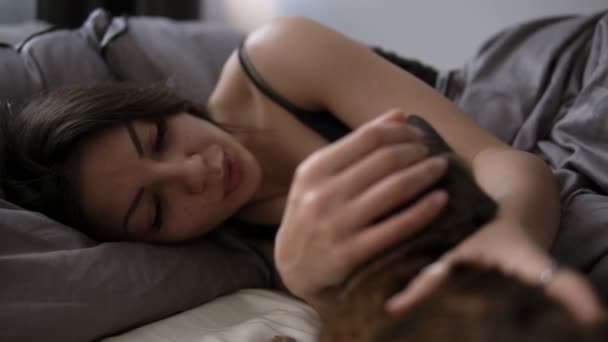 Zwierzęta i komfortu, reszta ludzi pojęcie - szczęśliwa młoda kobieta z kotem, leżąc w łóżku w domu. Brunetka dziewczyna miłej pieszczoty jej kota w łóżku. Widok z bliska — Wideo stockowe