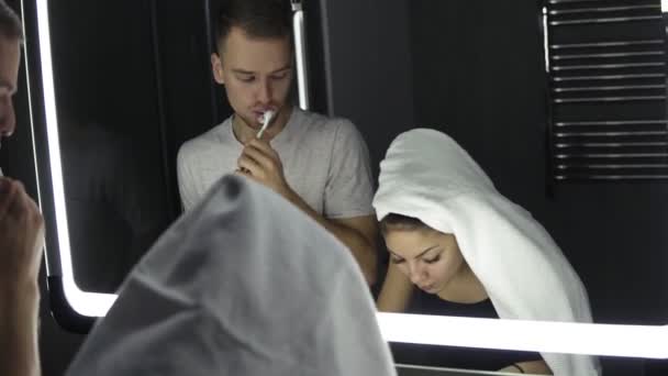 Joven pareja amorosa mirando en el espejo mientras se cepilla los dientes en el interior. Señora con toalla de baño en la cabeza. De cerca. — Vídeos de Stock