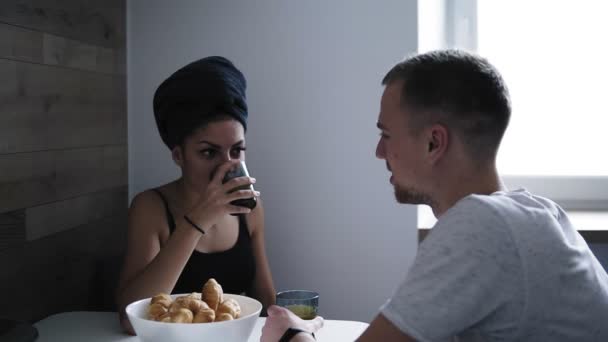 Schöne Frau nach der Dusche mit dunklem Handtuch auf dem Kopf, Croissants isst und Kaffee trinkt. Sie redet mit ihrem Freund vor sich hin und lacht. junges Paar frühstückt in der Küche — Stockvideo