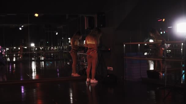 Zeldzame zicht op een sportieve, sexy meisje in korte broek en beha permanent in de hoek en het kiezen van de juiste muziek voor haar dansopleiding. Donkere, dim studio — Stockvideo