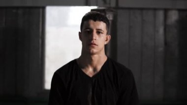 Kum torbası yakınındaki eski bir moda boks salonunda duran bir koyu saçlı beyaz erkek boxer portresi. Rahat kıyafetler ve siyah eldiven giyiyor. Terli ve yorgun eğitimden sonra