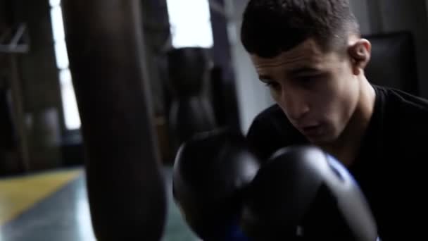 Imágenes de mano de boxeadores jóvenes entrenando. Hombre de pelo oscuro golpeando la bolsa de boxeo, patadas duras. La motivación en el deporte. Gimnasio de estilo antiguo, durante el día. De cerca. — Vídeos de Stock