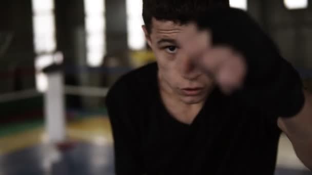 Giovane uomo compie colpi in bende da boxe nere e guardando seriamente la fotocamera in palestra. Da vicino stanco dal pugile allenamento. Boxe ring e palestra sullo sfondo. Rallentatore — Video Stock