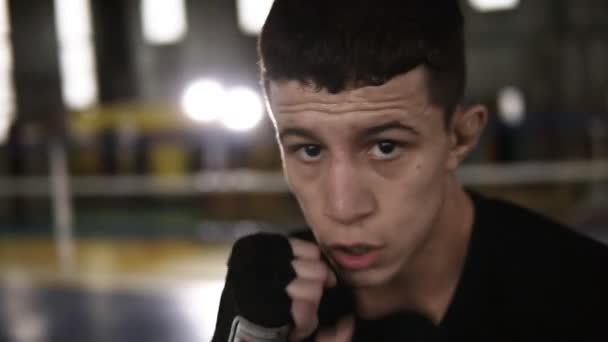 Hombre joven en camiseta negra cumple golpes en vendas de boxeo negro y mirando seriamente a la cámara en el gimnasio. Cansado del boxeador de entrenamiento. Anillo de boxeo, sofás y gimnasio en el fondo. Cierre — Vídeos de Stock