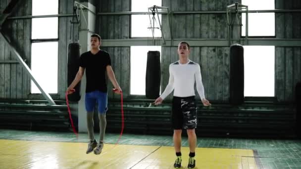 Jonge boksers in casual kleding springen met behulp van een springtouw tijdens de warming-up in boksen studio-gym. Hoge sprongen doen synchroon. Volledige lengte, slow-motion — Stockvideo