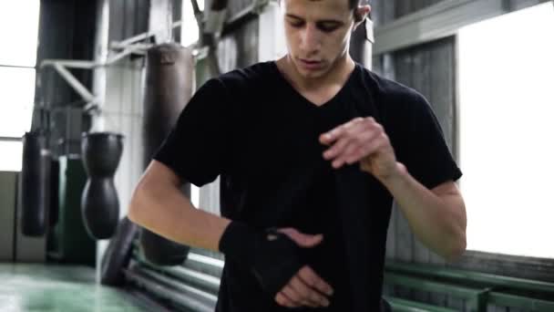 Jonge mannelijke bokser op zwart T-shirt staat in de oude stijl sportschool en wraps een hand zwarte pleister op zijn hand. Bokszakken op de achtergrond — Stockvideo