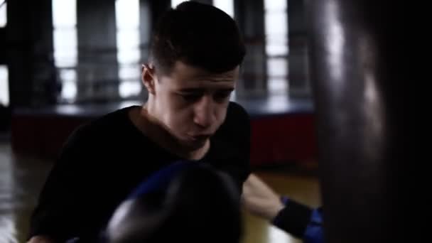 Hombre de pelo oscuro golpeando agresivamente la bolsa de boxeo, patadas duras. La motivación en el deporte. Gimnasio de estilo antiguo, durante el día. De cerca. — Vídeos de Stock