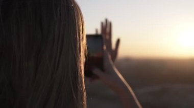 Uzun saçlı genç kadın arkadan ayakta bulanık şehir arka plan günbatımı ve gündoğumu, cep telefonu kullanır. Vaktinizi elini önünde fotoğrafını yapım üzerinde duran güneş ışınları