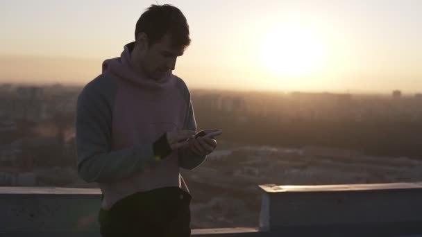 Hombre joven en sudadera con capucha rosa y negro usando teléfono inteligente al aire libre de pie en el techo. Paisaje urbano al atardecer o al amanecer — Vídeos de Stock