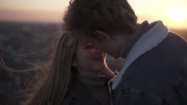 Glad, kärleksfull ungt par njuta av en romantisk kyss bakgrundsbelyst av solen med flare effekt stående på blåsiga taket med urban bakgrund — Stockvideo