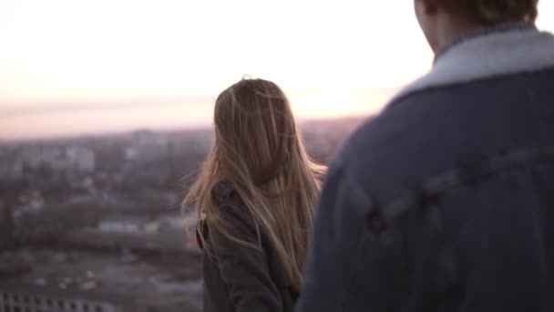 Millenial elegante pareja - chica de pelo largo y hombre rubio de pie en el techo ventoso abrazando con una hermosa escena de la salida del sol en el fondo. Paisaje urbano borroso alrededor — Vídeos de Stock