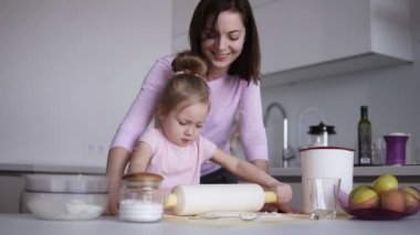 Sevinçle birlikte mutfakta yemek yaparken bir oklava ile hamur yayıyoruz sevimli küçük kızını yardım beyaz genç sevgi dolu bir anne. Ağır çekim