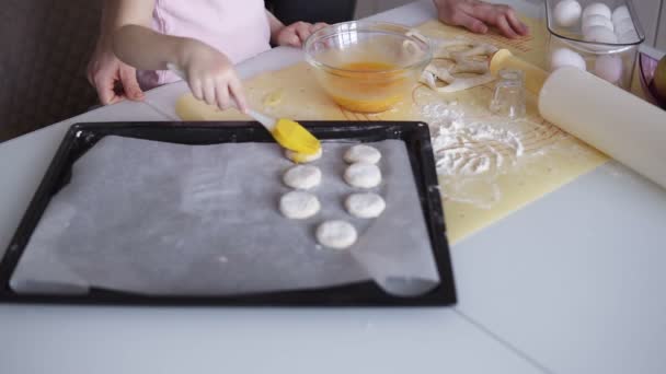 Concepto de familia, cocina y gente: la madre feliz y su hija pequeña en la cocina casera aplican galletas con huevos batidos antes de poner una bandeja de galletas para hornear. — Vídeo de stock