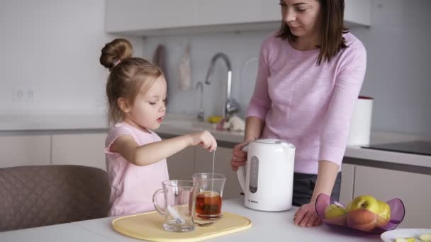 Hermosa madre y su niña en la cocina casera compartiendo momentos felices juntos. La morena mamá dejó que su pequeña niña hiciera una taza de té. Chica poniendo bolsa de té en taza transparente — Vídeo de stock