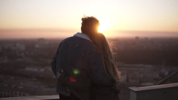 Joven mujer de pelo largo y hombre mirando a la ciudad al atardecer de pie en el techo del edificio alto. Observando la puesta de sol abrazándose. Vista trasera — Vídeo de stock