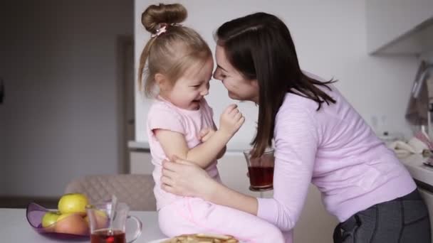 Morena mamá alegre jugando con su pequeña hija besándose, abrazándola y haciéndole cosquillas mientras la niña está sentada en la mesa en la cocina ligera. Buenos días, paternidad. De cerca. — Vídeos de Stock