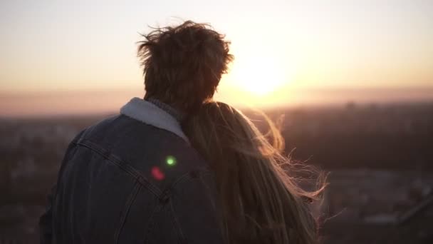 Gros plan de la jeune femme aux cheveux longs et de l'homme regardant la ville au coucher du soleil debout sur le toit du haut bâtiment. Regarder le coucher du soleil embrasser. Vue de derrière — Video
