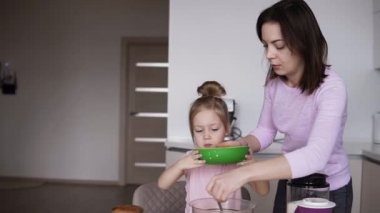 Genç anne ve kızı ev giysileri hamur krep için çok parlak bir mutfak yapmak. Sarı bir kase süt dökme. Modern mutfak. Ağır çekim