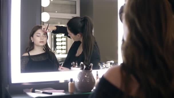 Make-up artiest met behulp van make-up borstel voor de toepassing van eyeshadows op ooglid fotomodel. Visagist make-up ogen doen met jonge vrouw in de schoonheidssalon. Spiegel reflectie beelden — Stockvideo