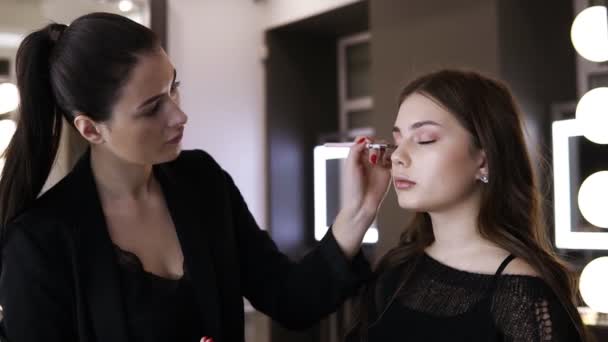 Bruneta žena nake-up umělce s červeným manikúra dělá dotyků eyeshadows na víčkách modelu. Světlý make-up studio s žárovky — Stock video