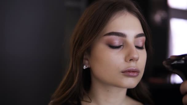 Retrato de una hermosa joven con maquillaje ligero y pelo largo y claro en el estudio de maquillaje. Visagista corrigiendo la superficie de una piel usando un aerógrafo — Vídeo de stock