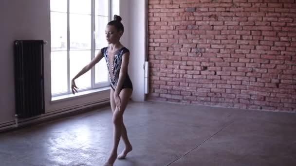 Hezká dívka tančí rytmické gymnastiky v umělecké kombinézu v blízkosti oken. Mladá gymnastka cvičení v gymnastické školy nebo studio s červenou cihlovou zeď na pozadí — Stock video