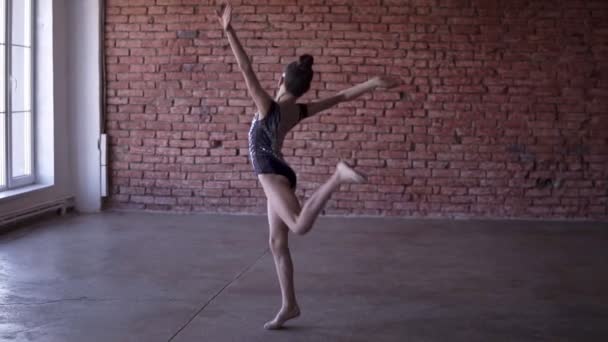 Schattig gymnast doen spinnen in onze ontwerpstudio baksteen op muur en venster achtergrond. Turnen meisje gekleed zwarte en gekleurde Romper. Slow motion — Stockvideo